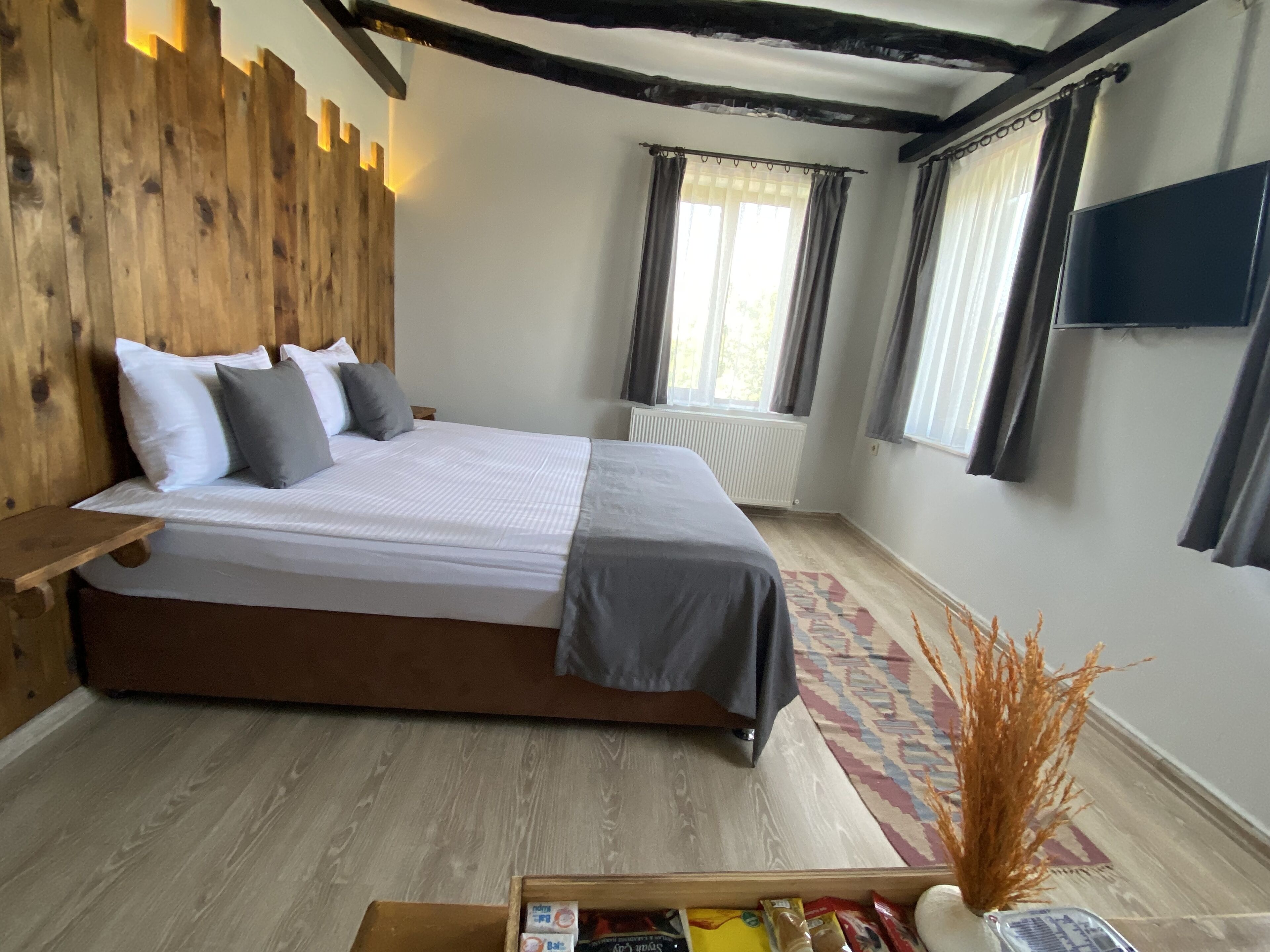 Un dormitorio con cabecera de madera, una cama grande con ropa de cama gris, una mesita de noche, un radiador, una ventana con cortinas, un televisor montado en la pared y una pequeña mesa con objetos encima.