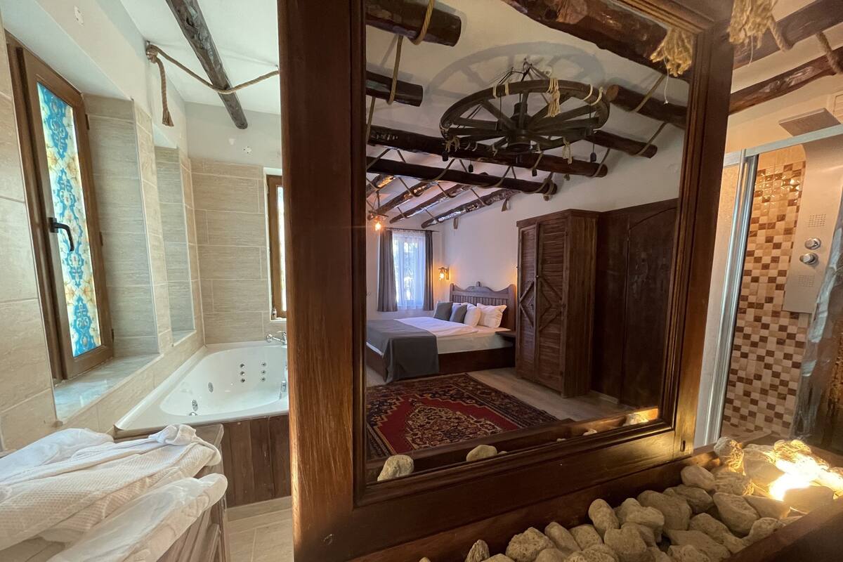 A bathroom with a large bathtub, a mirror reflecting a bedroom with a bed and a rug, and a stone wall.