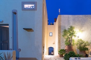 A narrow street with white walls and a blue sky. There are potted plants and a door on the left.