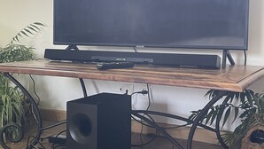 Un téléviseur à écran plat posé sur un meuble en bois, avec un haut-parleur en dessous, dans une pièce dont le sol est carrelé et où se trouvent des plantes en pot.