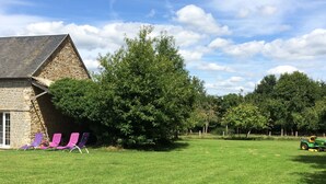 Un bâtiment en pierre avec un toit en pente, une cour gazonnée et un grand arbre.