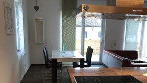 A modern kitchen with a built-in stove, a dining area with a table and chairs, and a view through large windows. - Schön Apartments (Ohmden)