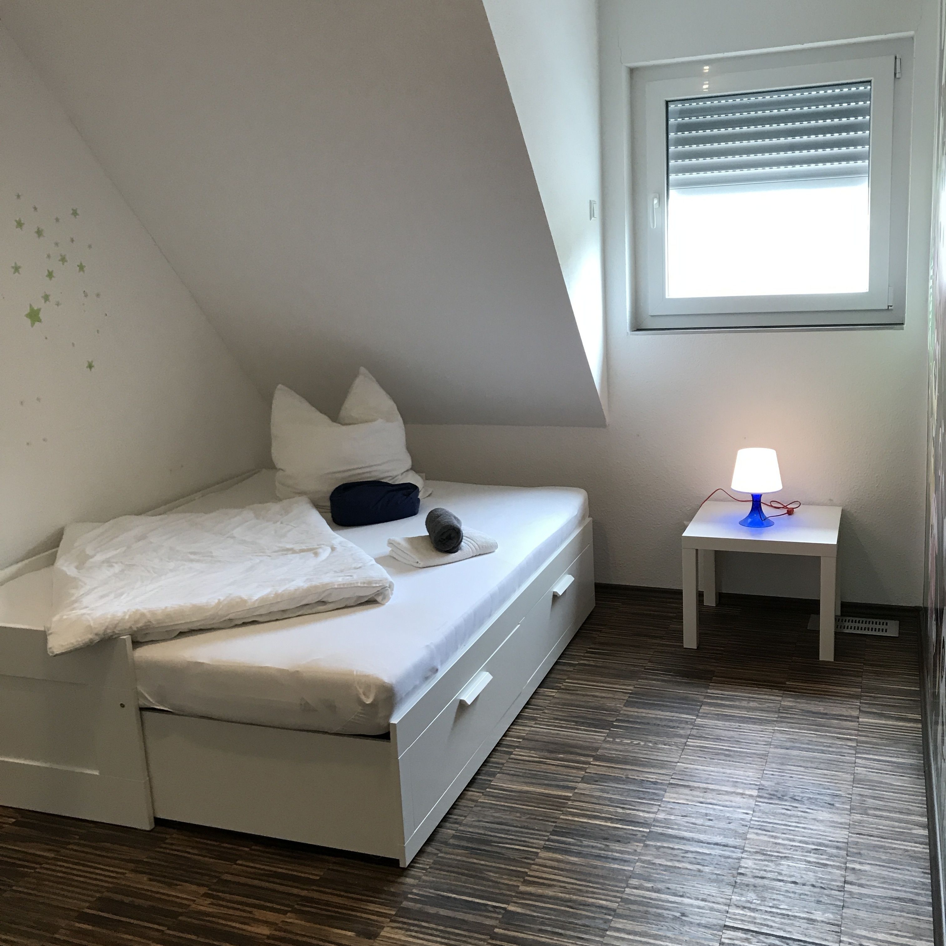 A small, neatly arranged bedroom with a bed, a bedside table with a lamp, and a window with blinds.