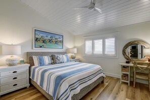 A bedroom with a bed, two bedside tables, a ceiling fan, a window with blinds, a wall-mounted mirror, and a painting above the bed.