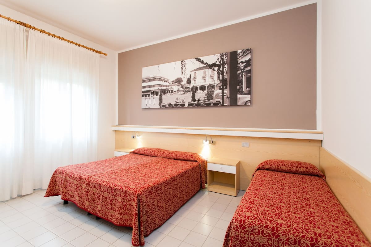 A hotel room with two single beds, a large window with sheer curtains, a wall-mounted picture, and a bedside table with a lamp.