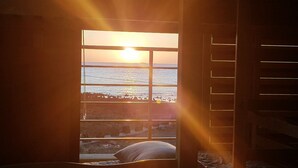 A room with a large window overlooking a beach at sunset.