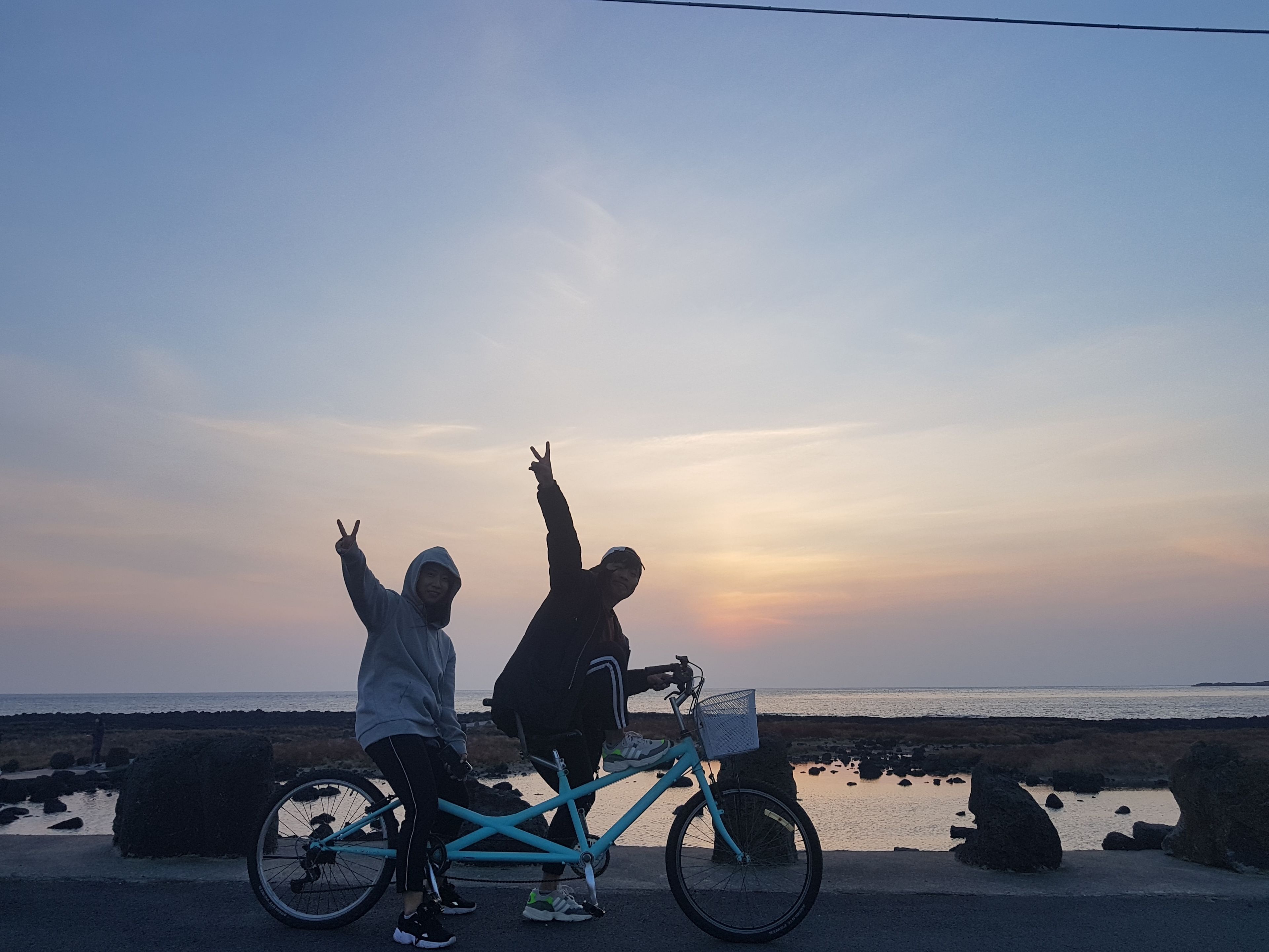 Zwei Personen fahren mit einem Fahrrad an einer Küstenstraße, im Hintergrund ein Sonnenuntergang.