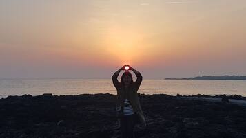 A person standing on a rocky beach with the sun setting in the background.