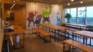 A modern dining area with wooden tables and benches, a mural of superheroes on the wall, and large windows offering a view.
