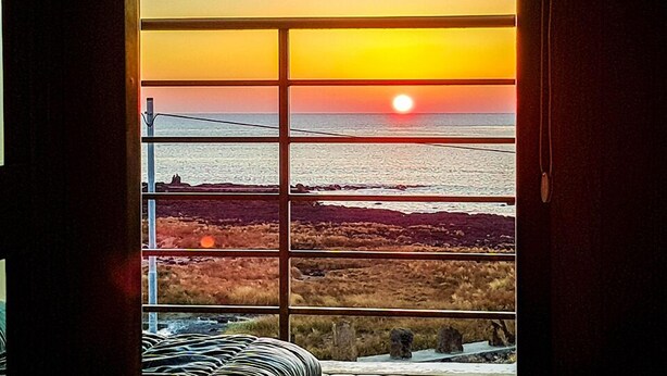 A view through a window with a sunset over the ocean, a sandy beach, and a rocky shoreline.