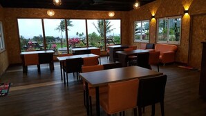 Un ristorante con tavoli di legno e sedie arancioni, ampie finestre che offrono vista su spazi verdi e palme, e luci calde.