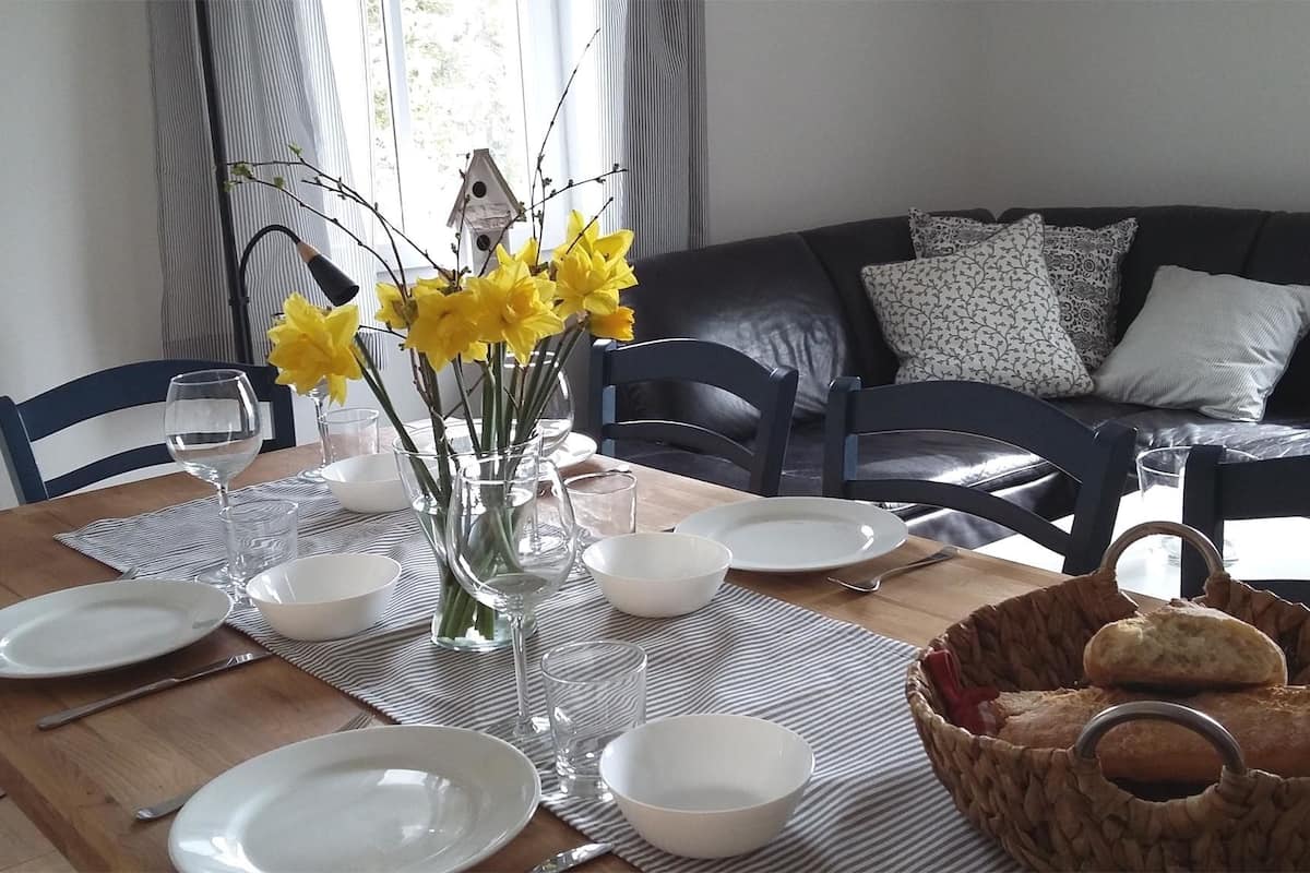 Une table à manger est dressée avec des assiettes, des bols et un vase rempli de fleurs jaunes. Un panier de pain est posé sur la table.