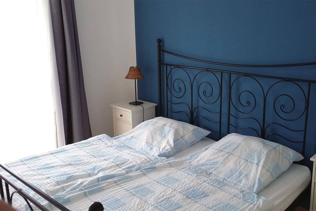 Un lit avec un cadre en métal, une literie blanche, une table de chevet avec une lampe et un mur avec un détail bleu.