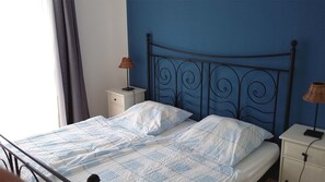Un llit amb estructura metàl·lica, roba de llit blanca, una tauleta de nit amb una làmpada i una paret amb detall blau.