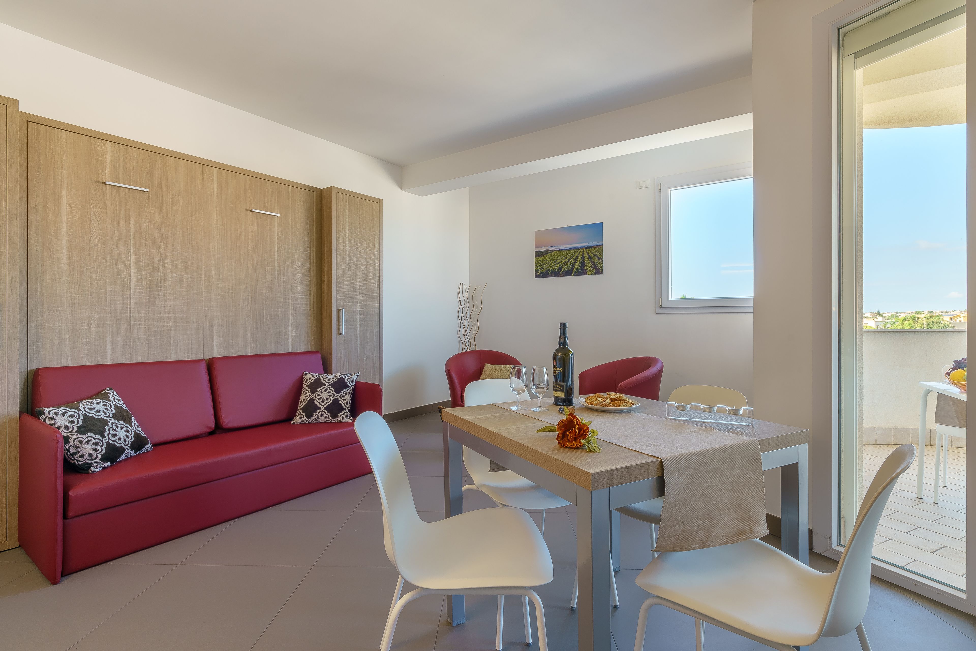 A modern living room with a red sofa, white chairs, a wooden cabinet, and a dining table with a bottle and glasses.