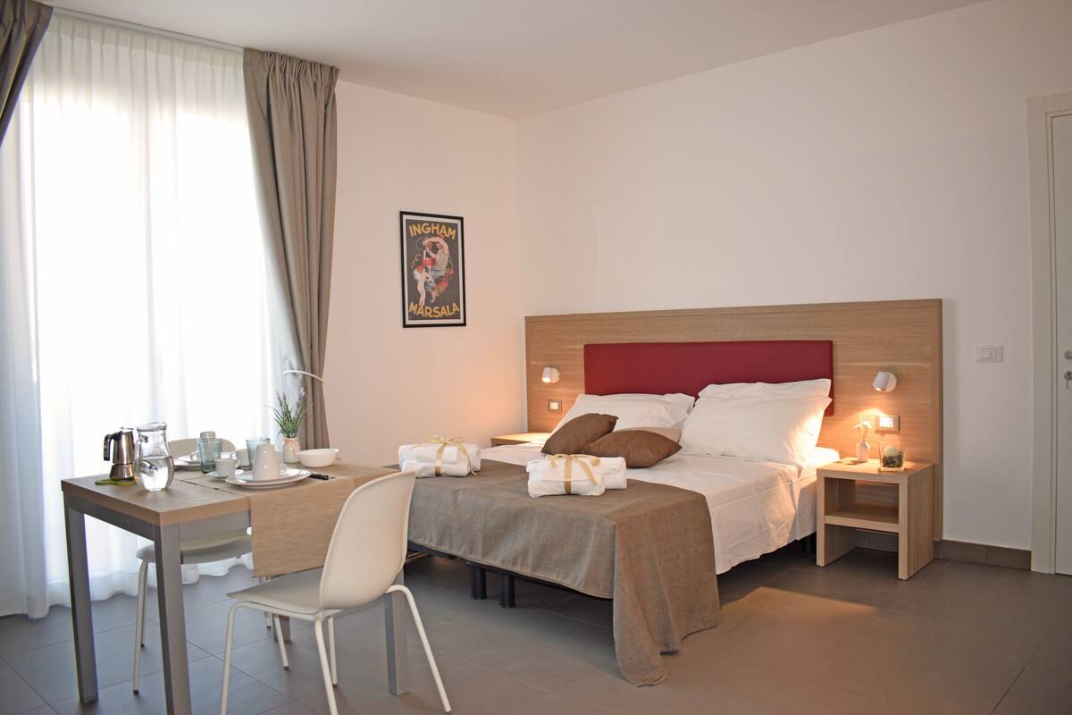 A hotel room with a bed, bedside table, chair, and a small table with a pitcher, cups, and a bowl.