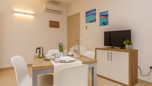 A modern dining area with a wooden table, white chairs, a television on a cabinet, and a wall-mounted air conditioner. - Travini Hotel Residence (Marsala)
