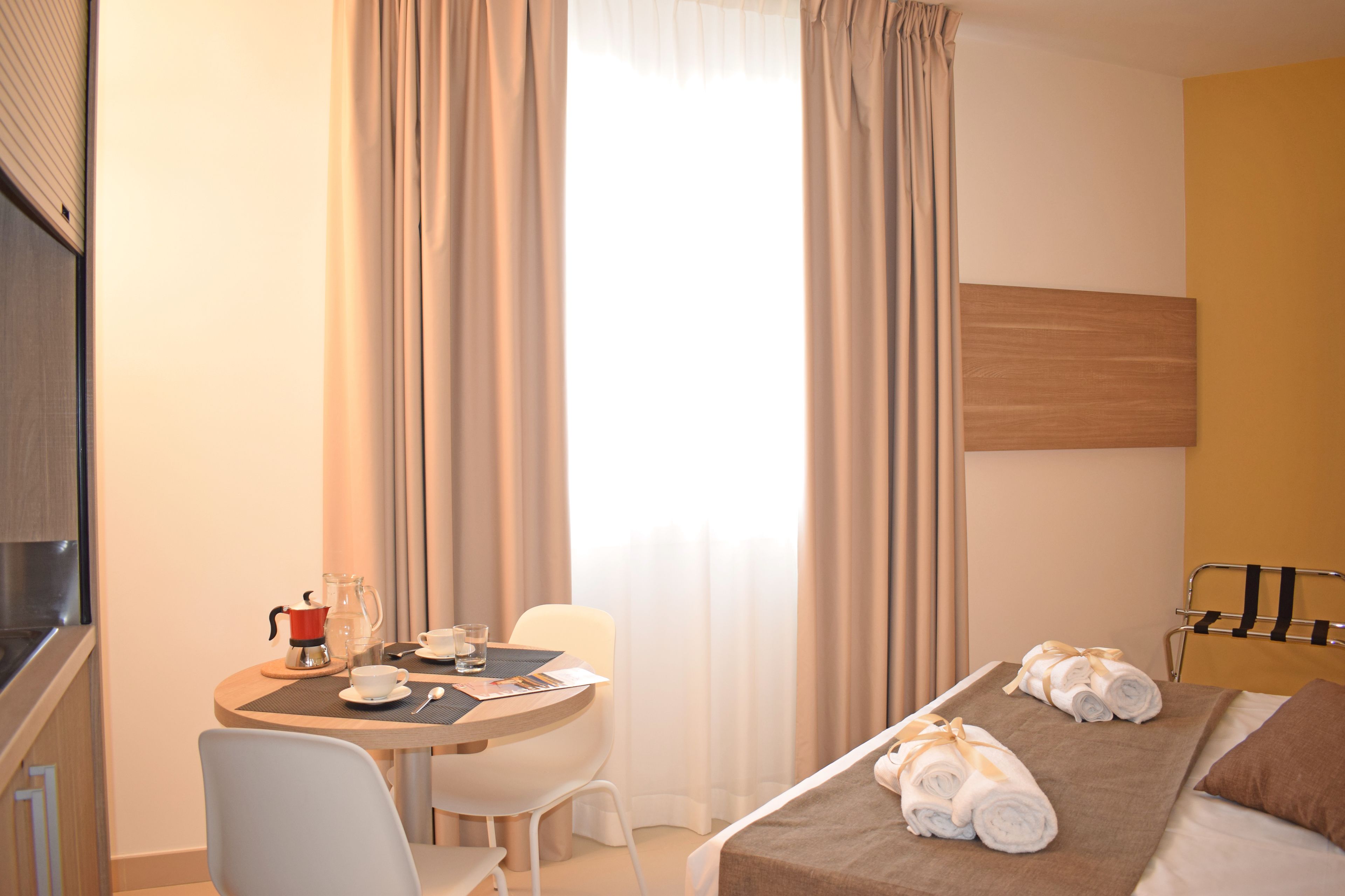 Habitació d'hotel amb llit, escriptori amb cafetera, cadira i finestra amb cortines.