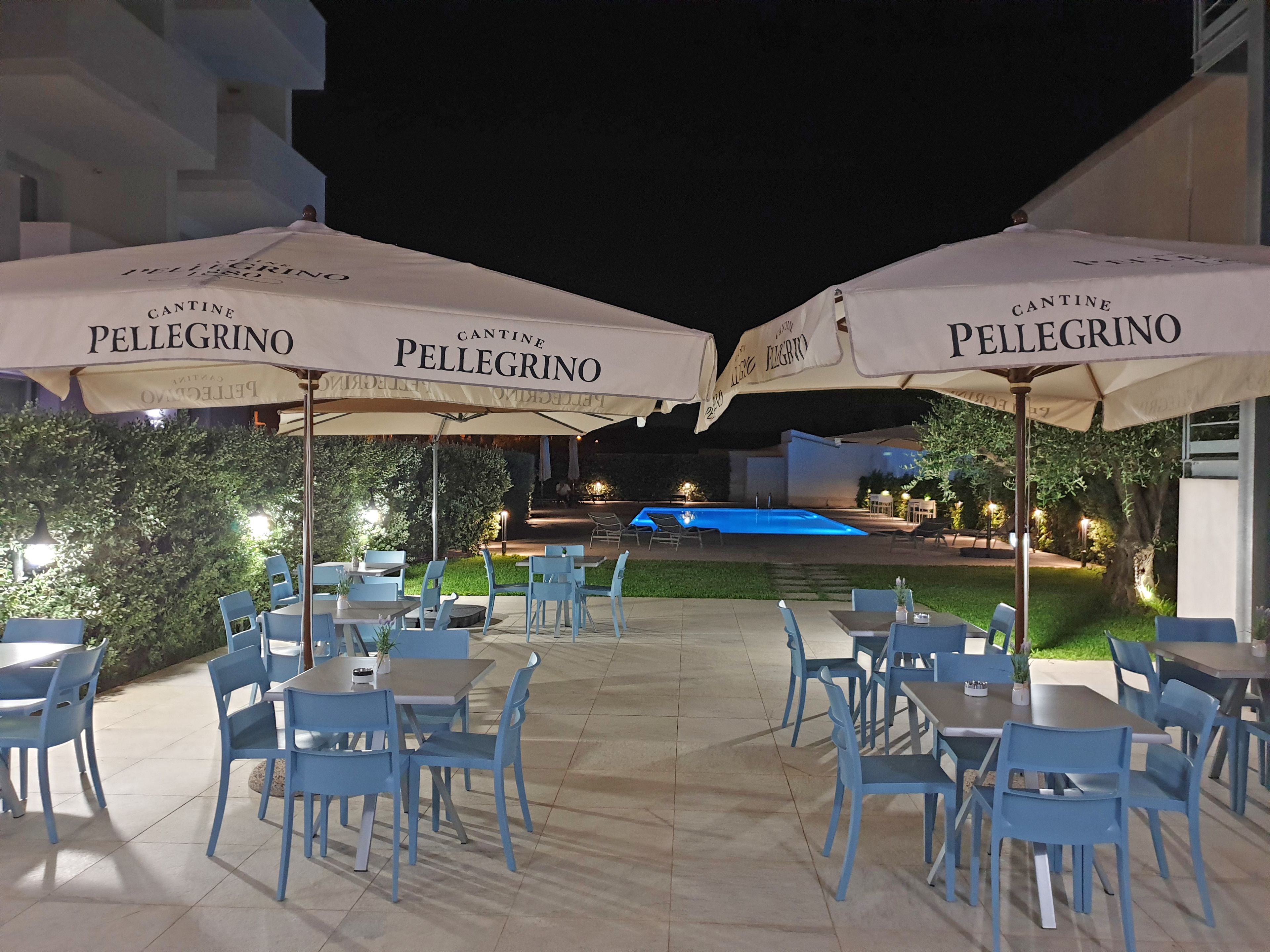 An outdoor dining area with blue chairs and tables under white umbrellas with 'Cantine Pellegrino' branding. A swimming pool is visible in the background.