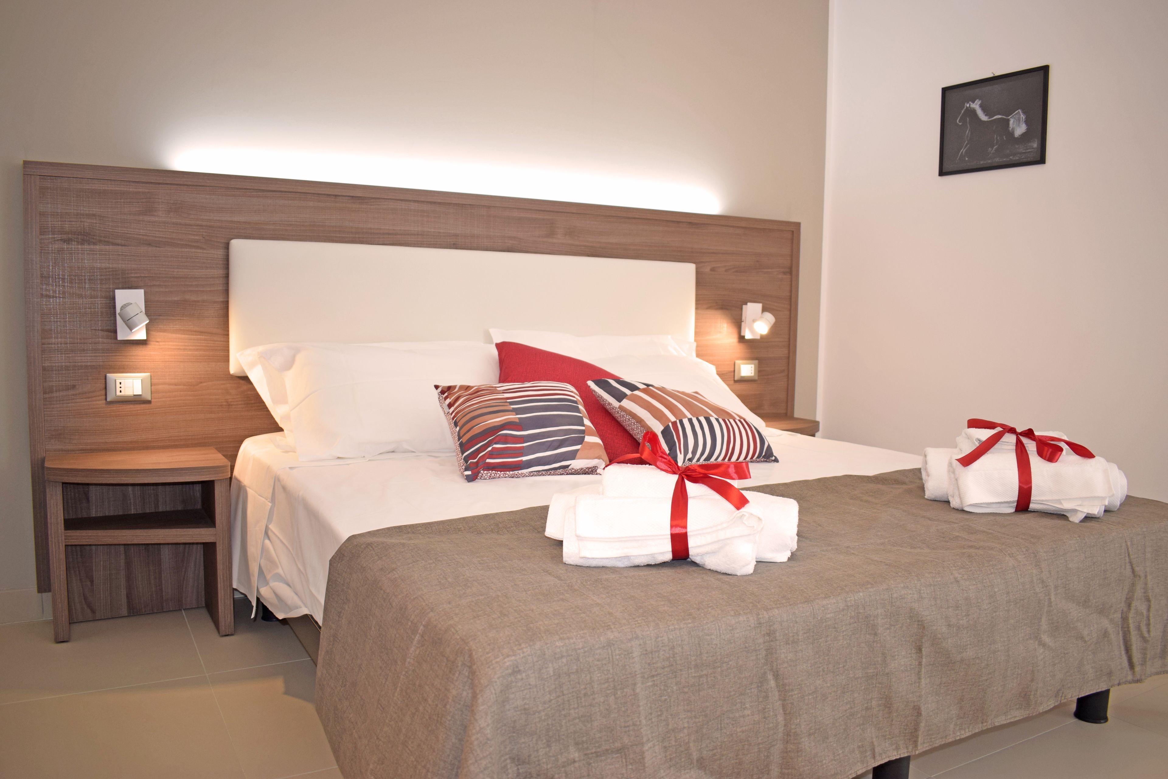 Una cama bien hecha con cabecera de madera, ropa de cama blanca y un cojín a rayas rojas y blancas. Hay dos mesitas de noche con lámparas, y un cuadro enmarcado en la pared.