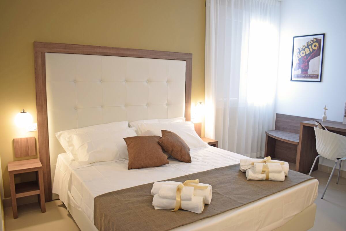 A neatly made bed with a wooden headboard, two bedside tables with lamps, a desk with a chair, and a framed picture on the wall.