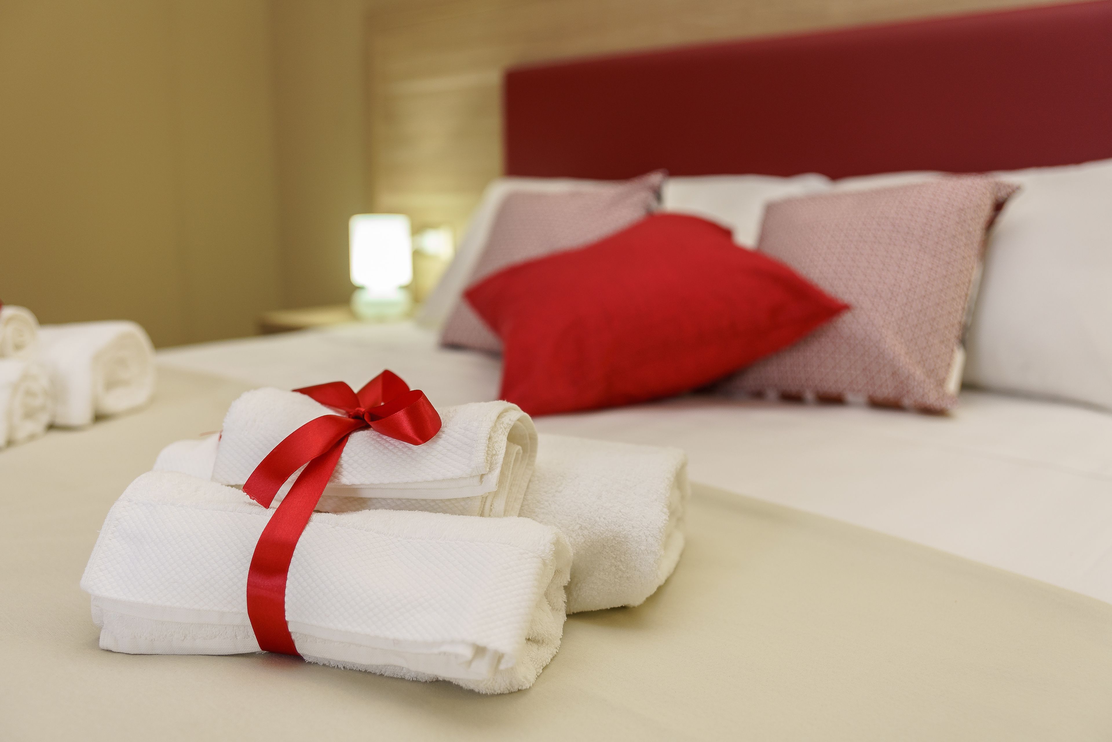 A neatly made bed with white linens and a red pillow, and a stack of folded towels with a red ribbon.