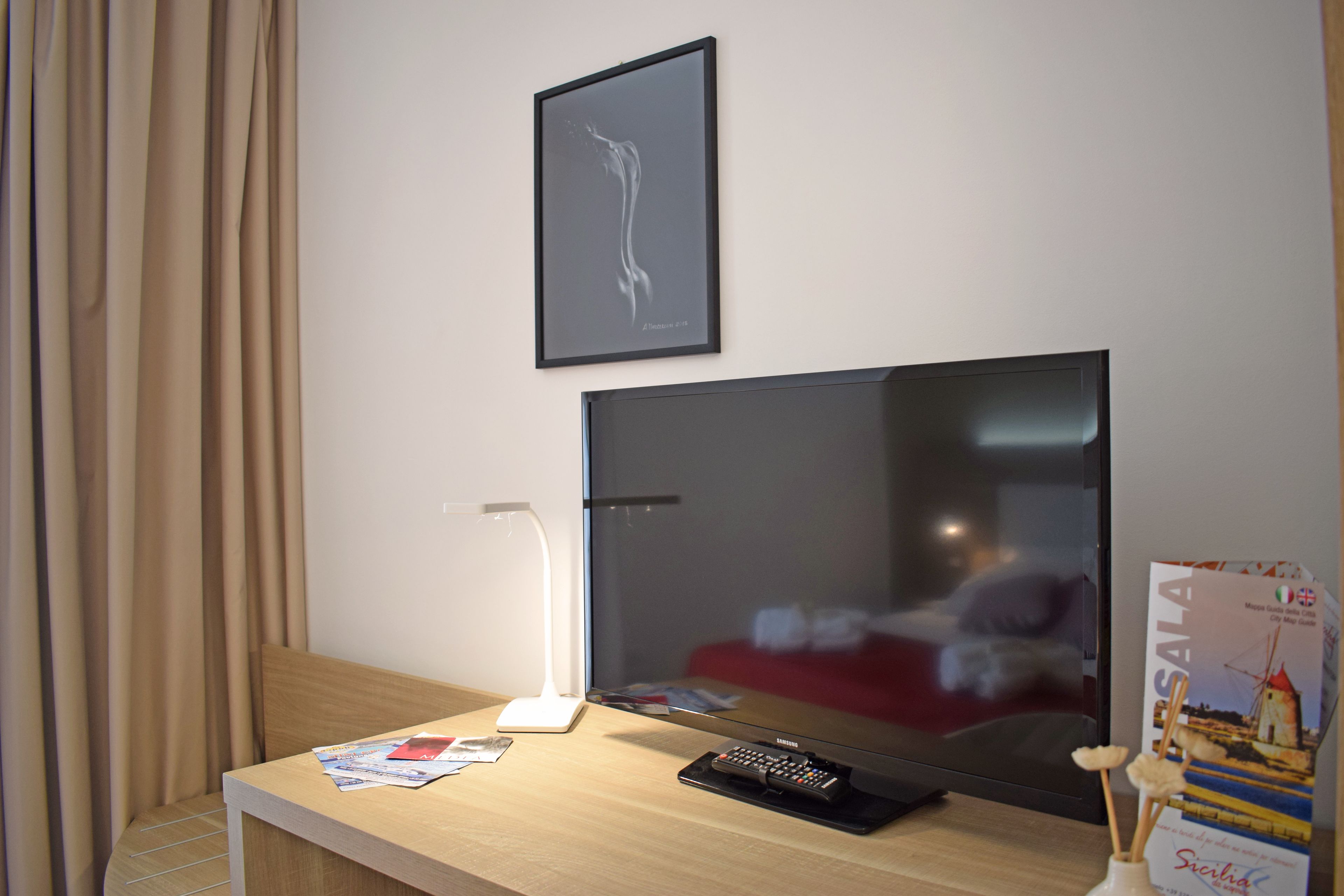 Una habitación de hotel con un televisor de pantalla plana grande, una mesita de noche con lámpara, un control remoto y un cuadro en la pared.