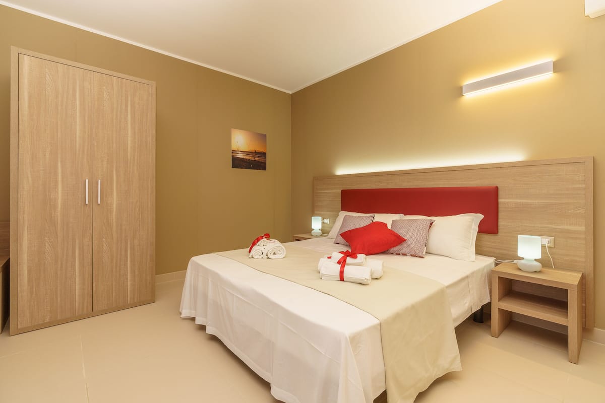 A hotel room with a large wardrobe, a bed with white bedding and red pillows, a bedside table with a lamp, and a wall-mounted light fixture.