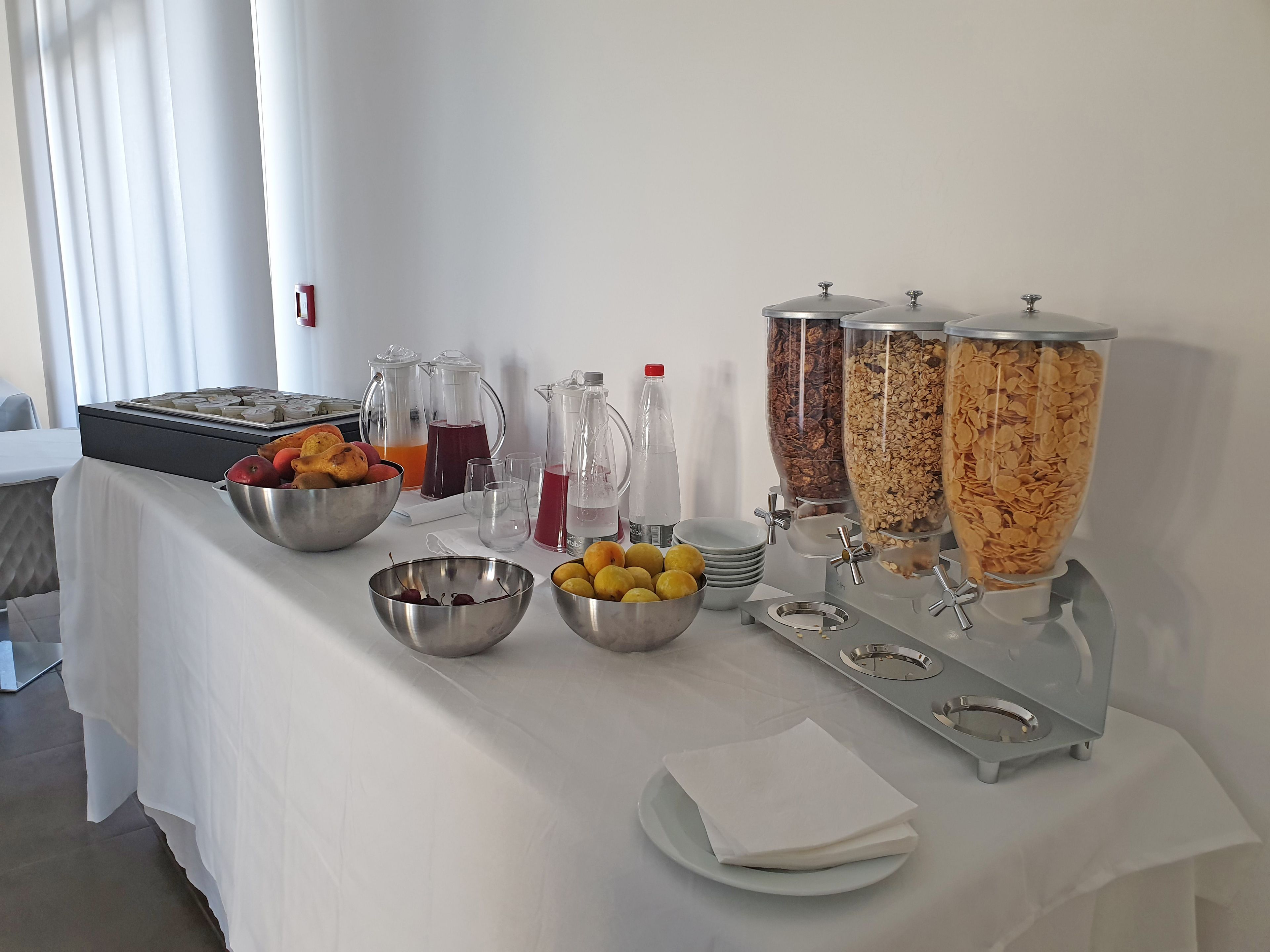 A buffet table with various food and drink options, including bowls of fruits, pitchers of beverages, and dispensers of cereal and granola.