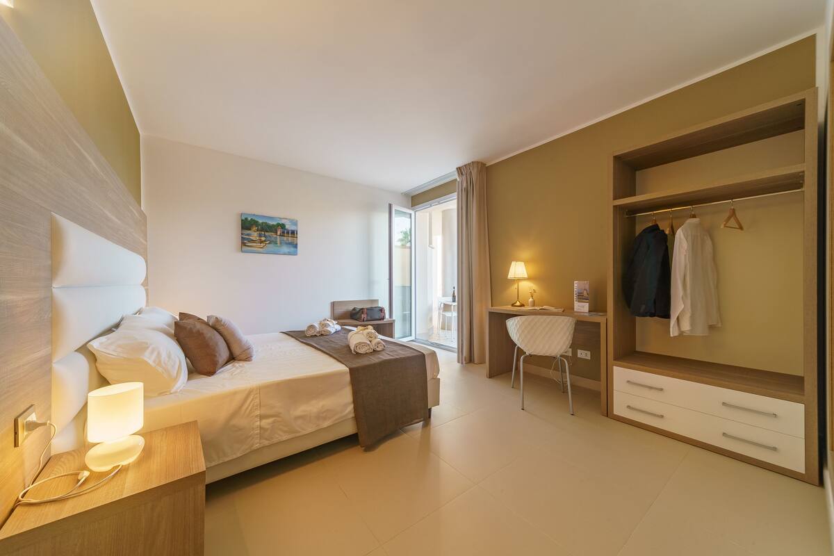 A modern hotel room with a bed, bedside table, desk, chair, and wardrobe.