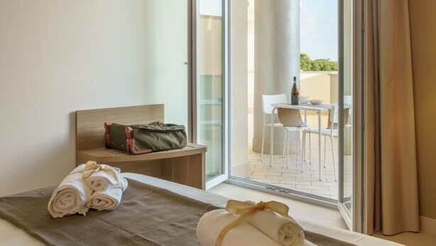 A hotel room with a bed, a bench with a bag, a table with chairs, and a view of the outside. - Travini Hotel Residence (Marsala)