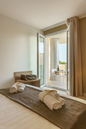A hotel room with a bed, a bench with a bag, a table with chairs, and a view of the outside. - Travini Hotel Residence (Marsala)
