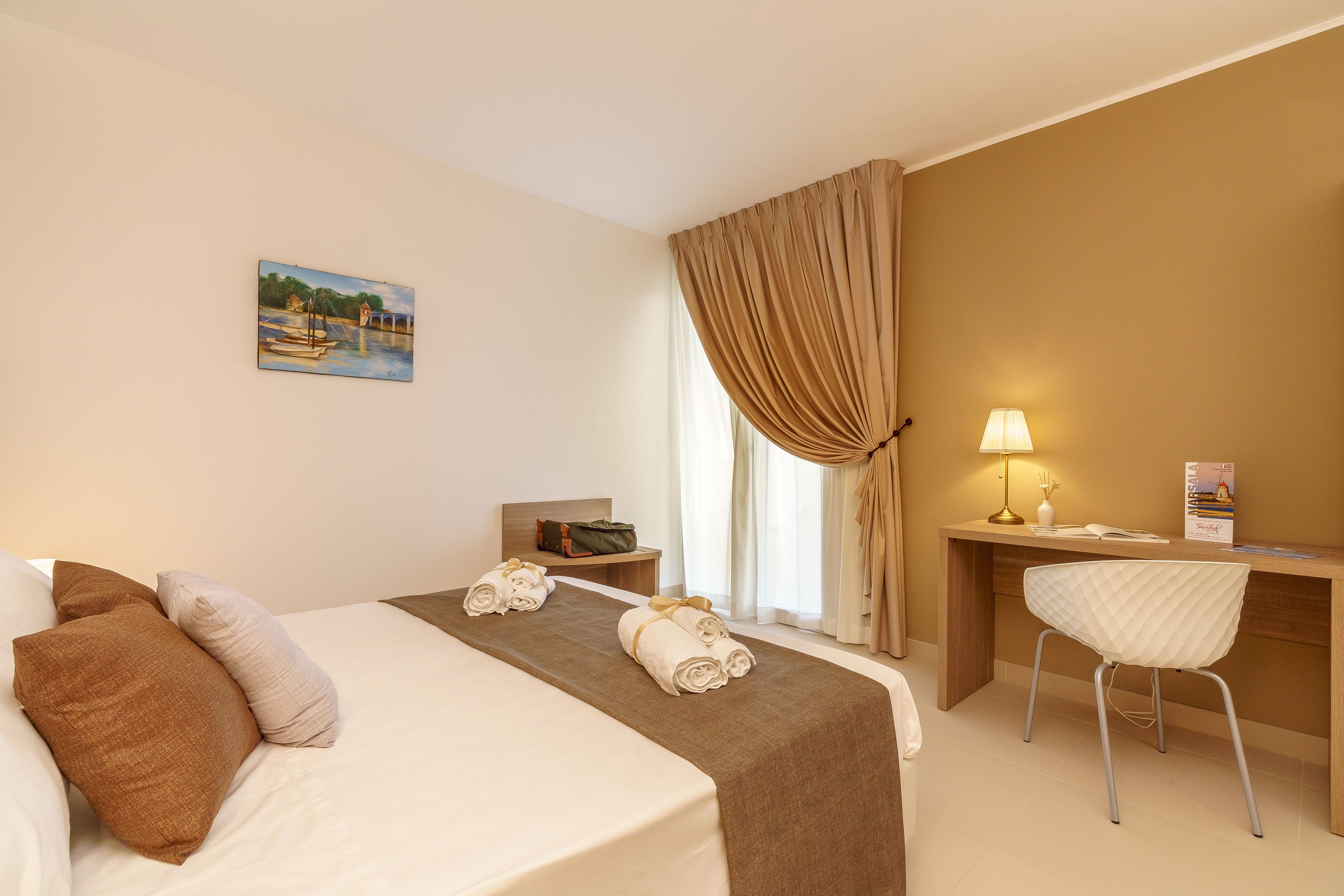 A hotel room with a bed, a desk, a chair, a lamp, and a painting on the wall.
