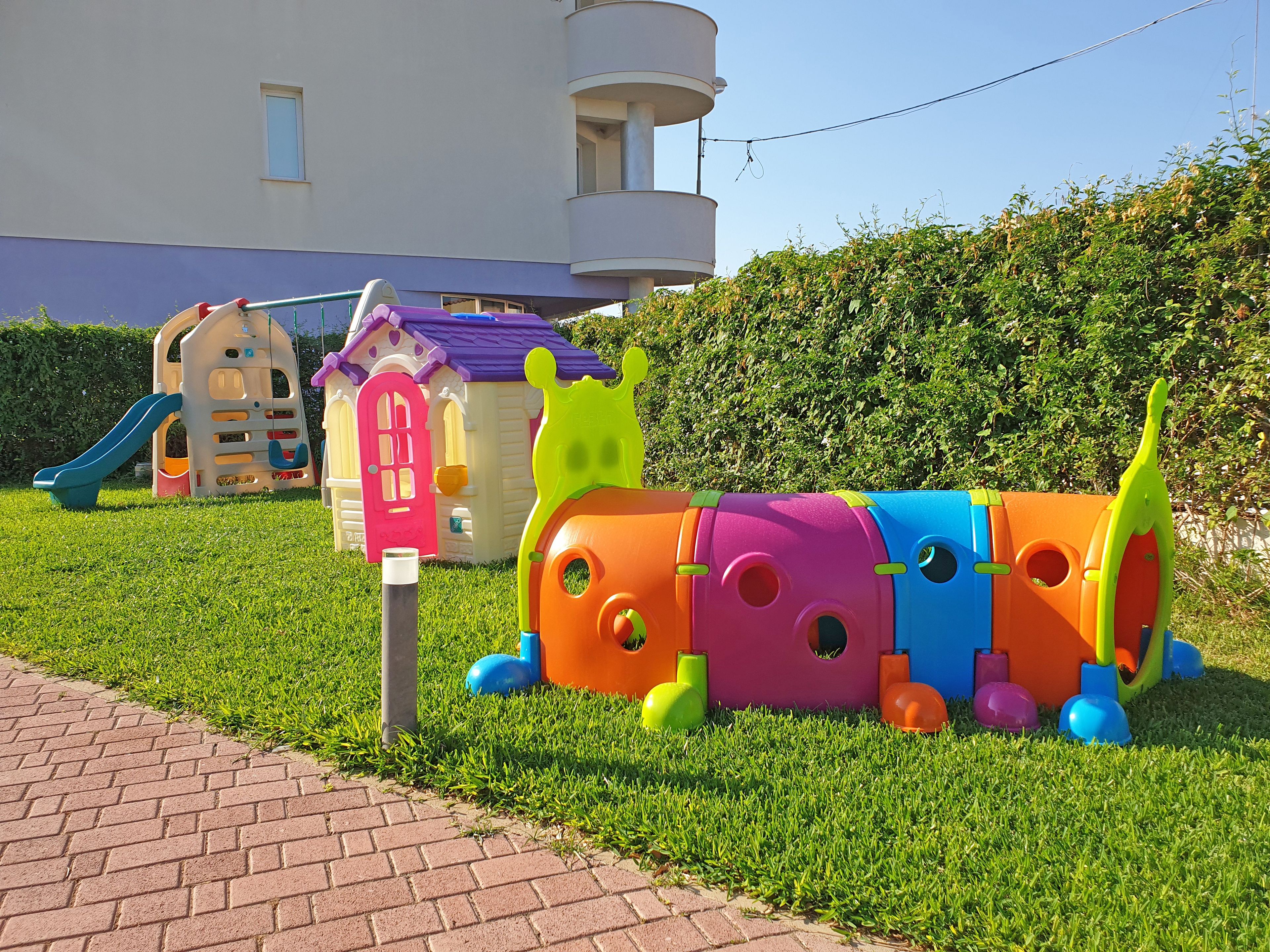 A children's play area with colorful play structures, a brick pathway, and a building in the background.