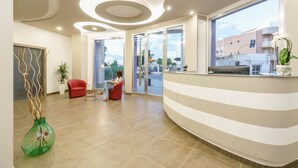 A modern reception area with a curved reception desk, a red chair, a potted plant, and a view of the outside through large windows. - Travini Hotel Residence (Marsala)
