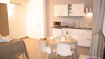 A hotel room with a bed, a dining table with chairs, a kitchen area with cabinets and appliances, and a bathroom with a shower curtain.