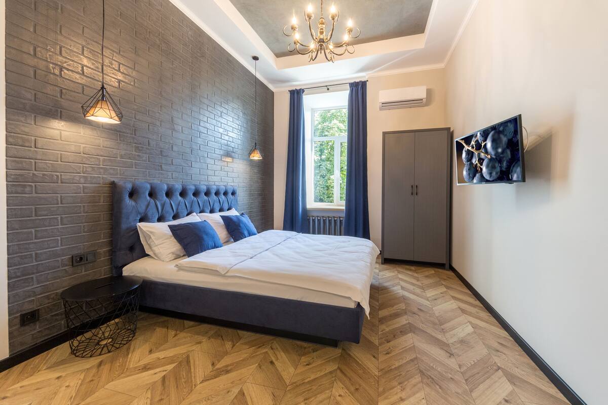 A modern bedroom with a brick wall, a tufted headboard, a large bed, a TV on the wall, and a window with curtains.