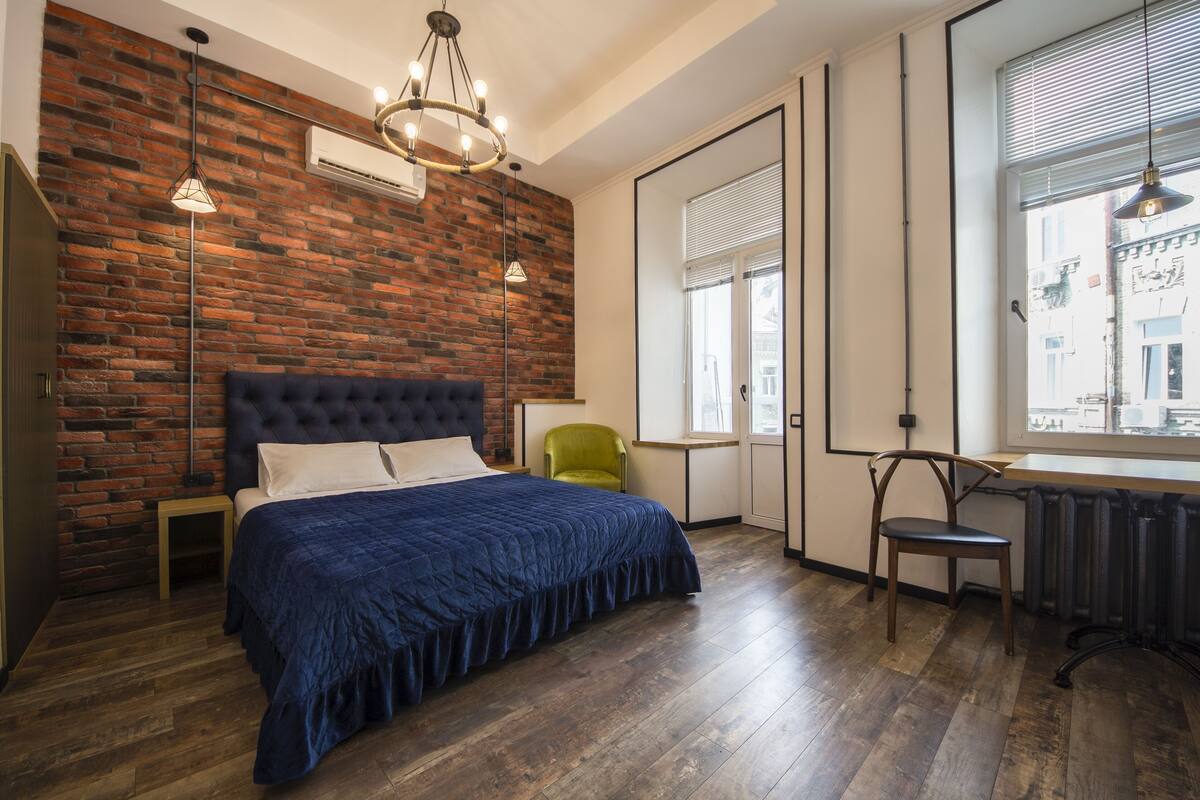 A bedroom with a brick wall, a large bed, a green chair, a wooden table, and a window with blinds.