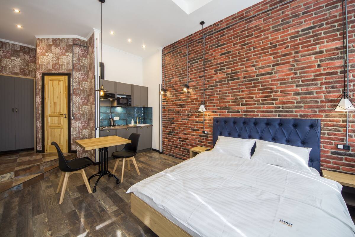A modern bedroom with a large bed, a dining area with a table and chairs, a kitchen, and a brick wall.
