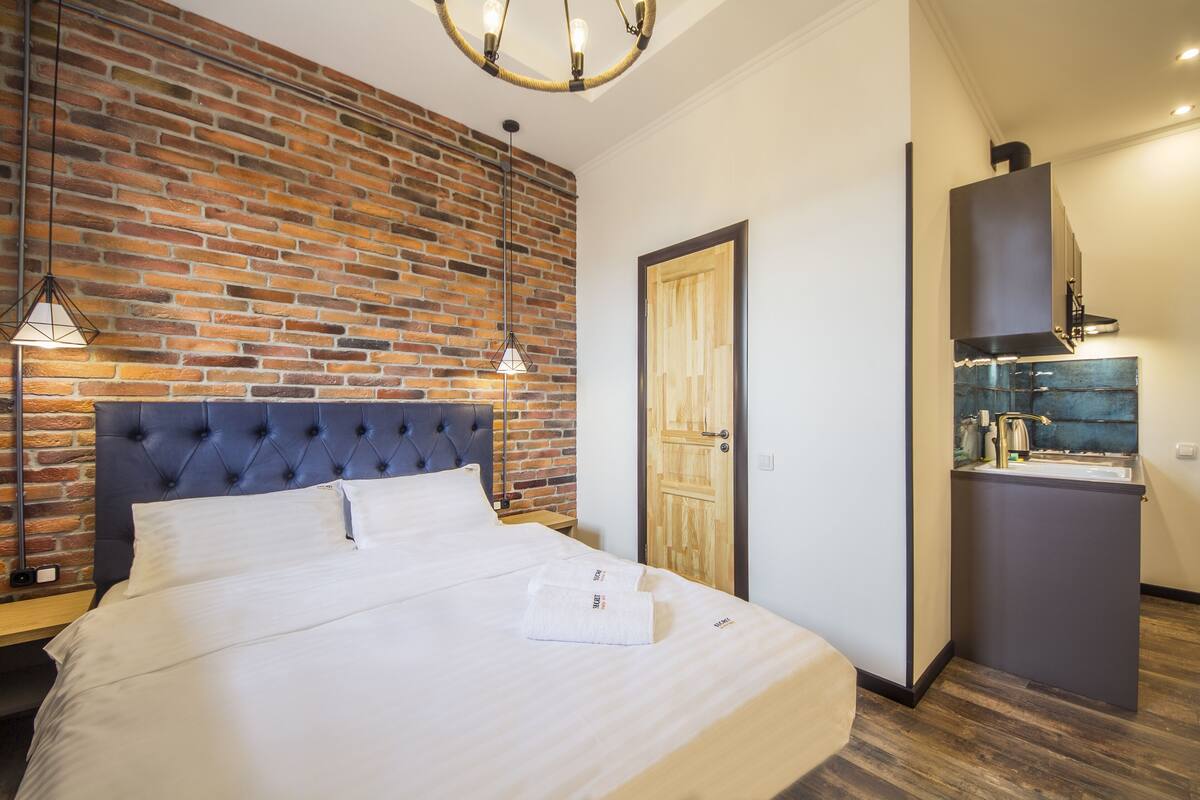 A hotel room with a brick wall, a tufted headboard, a wooden door, and a kitchenette.