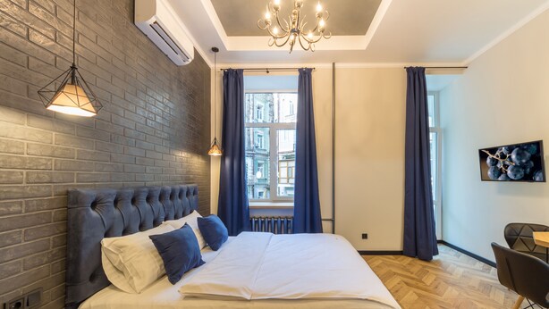 Quarto moderno com parede de tijolos, uma cama grande com roupa de cama branca e azul, um candelabro e vista para edifícios através de uma janela.