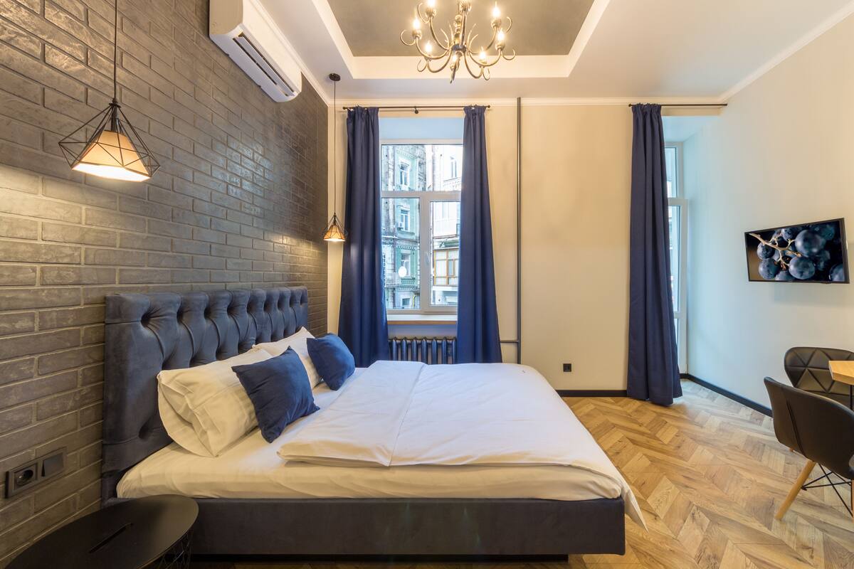 A modern bedroom with a brick wall, a large bed with white and blue bedding, a chandelier, and a view of buildings through a window.