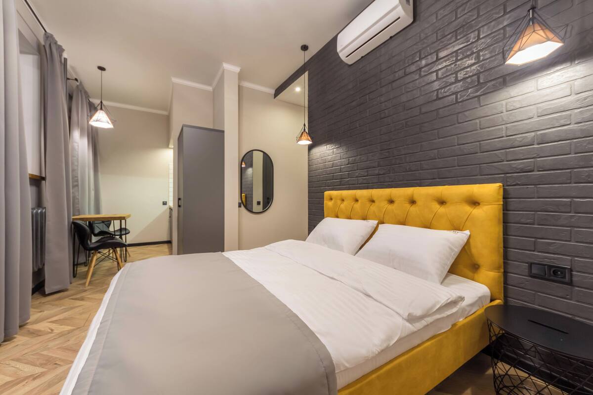 A modern bedroom with a yellow tufted headboard, a large bed with white linens, a wooden floor, a small table with chairs, and a black brick wall.
