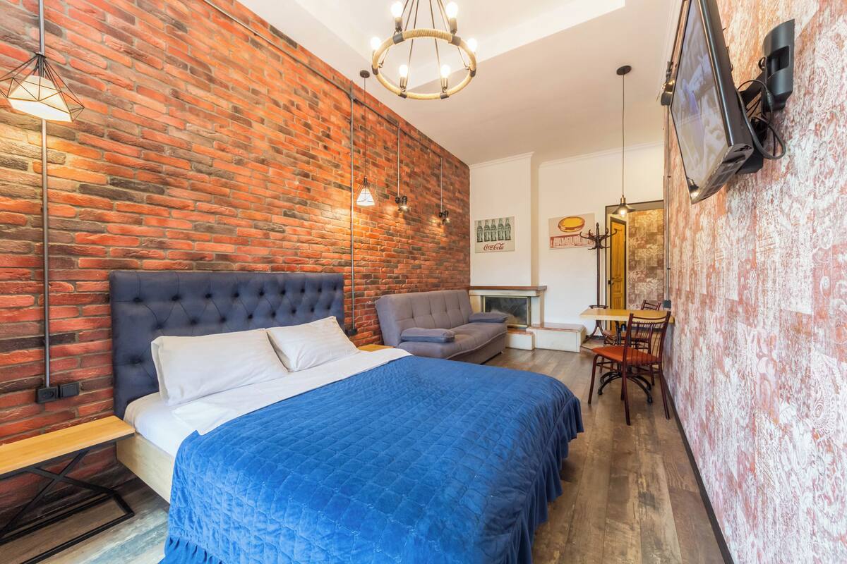 A bedroom with a brick wall, a tufted headboard bed, a blue bedspread, a grey sofa, a wooden table, and a TV mounted on the wall.