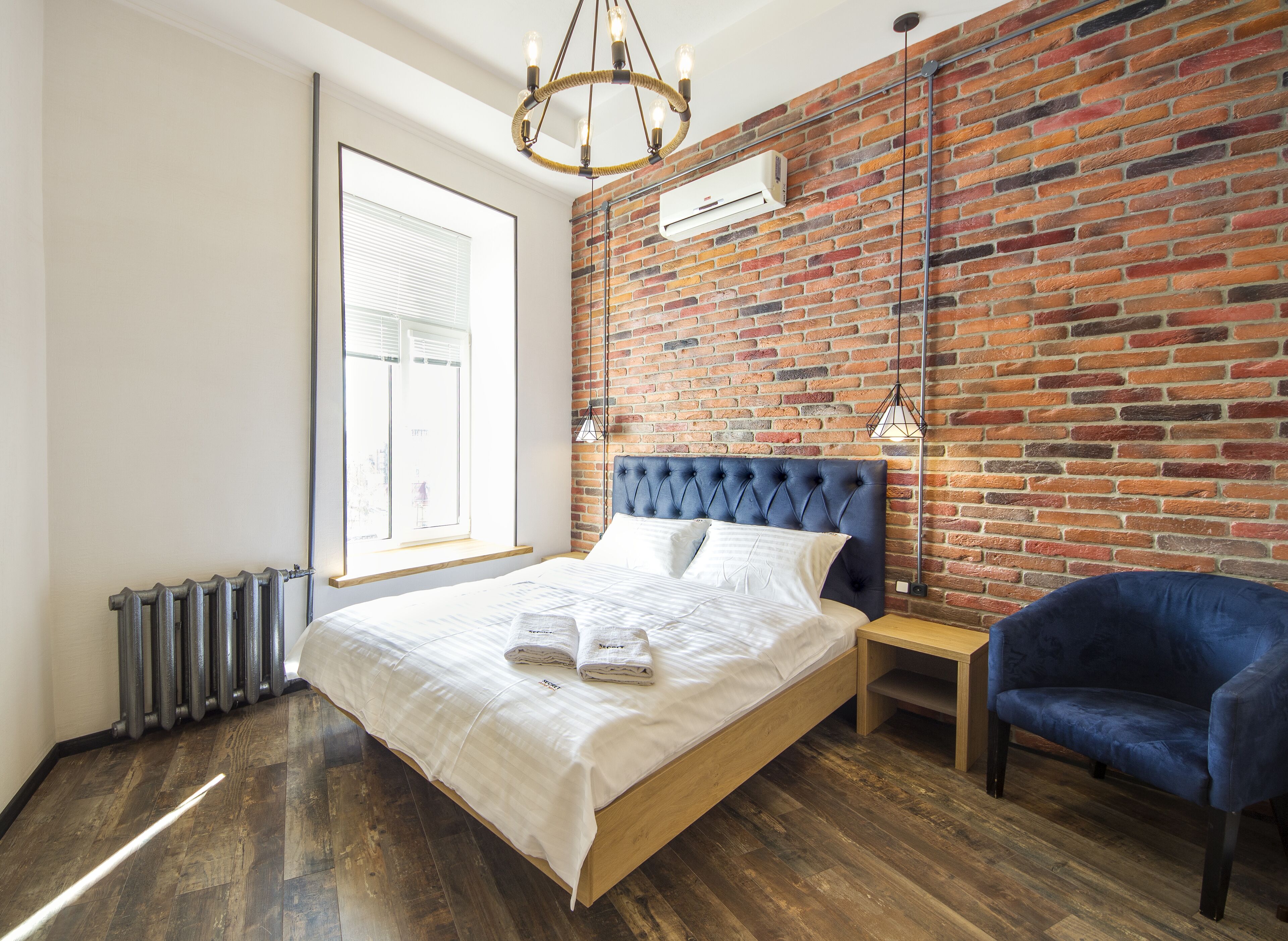 Un dormitorio moderno con pared de ladrillo, una cama grande, un sillón azul y una lámpara colgante.