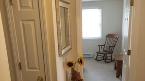 A hallway with a white door, a small white console table with a vase of sunflowers, a rocking chair, and a carpeted floor.