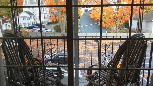 A view through a window with a metal railing, featuring a wooden deck and chairs.