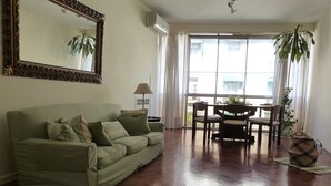 Ein Wohnzimmer mit Sofa, einem Esstisch mit Stühlen, einem großen Fenster mit Vorhängen und einer Zimmerpflanze.