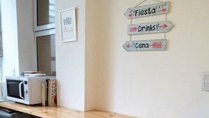 Eine Küche mit einer Holztresen, zwei Barhocker, einer Mikrowelle und einem Kühlschrank. An den Wänden befinden sich Schilder mit Hinweisen zu 'Fiesta', 'Getränke' und 'Cena'.