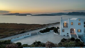 A white building with a balcony, a paved area, and a view of the sea and islands.